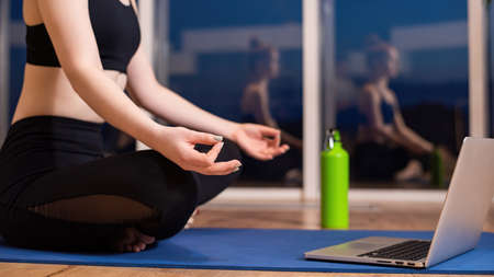 Young woman in sportswear is meditating on a yoga mat with laptop in front of her. Windows, duskの写真素材