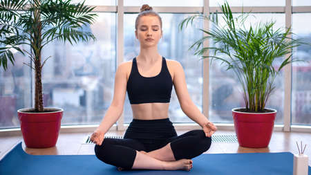 Young blonde woman in sportswear is meditating on a yoga mat with closed eyes. Bright room with...