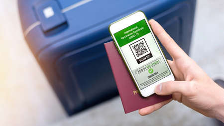 Male hand holding passport and smartphone with International Vaccination Certificate COVID-19 QR code, suitcaseの写真素材