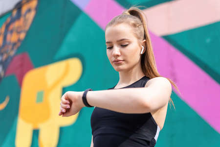 Young blonde woman at outdoors training in sportswear is on her fitness bracelet, multicolored...