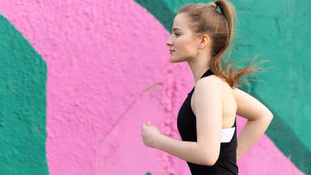 Young blonde woman in sportswear running on the road at outdoors training, multicolored wall on the...