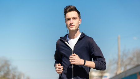 Young focused man running with earphones and fitness bracelet, jacket. Sunny weather