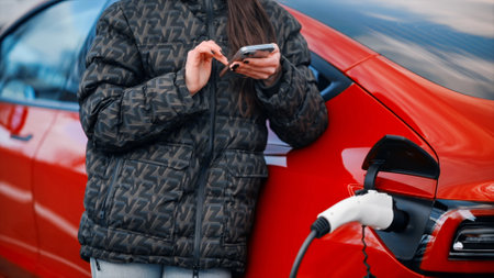 Close view of a woman using smartphone near her charging red electric carの写真素材
