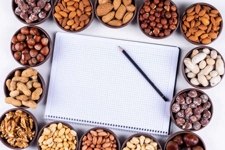 High angle view assorted nuts and dried fruits in rectangle shaped mini different bowls with pecan, pistachios, notebook with pen on white wooden background. horizontal space for textの写真素材