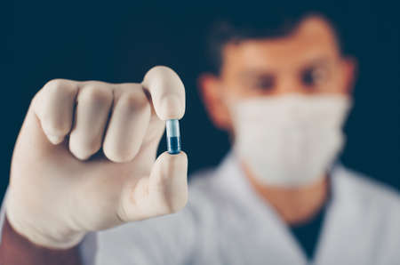 Close-up doctor with mask and gloves holding blue pill with his fingers on dark background. horizontalの写真素材