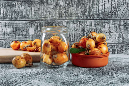 Some loquats in a jar, bowl and cutting board on gray textured background, side view.の写真素材