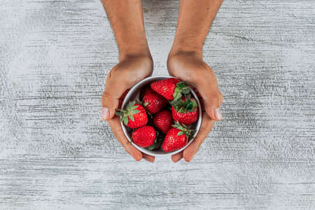 Man holding strawberries in a bowl on light wooden background, top view.の写真素材