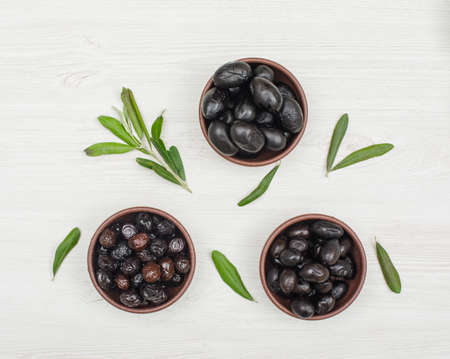 Black and brown olives in a clay bowls with olive tree branch and leaves top view on a white wood backgroundの写真素材