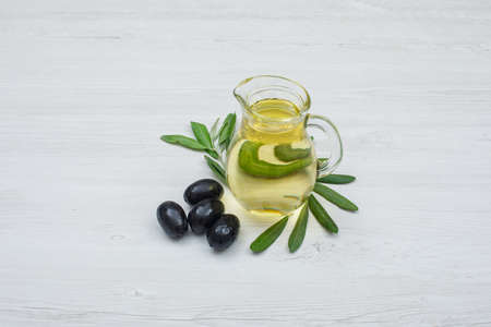 Black olives and olive oil in a glass jar with olive leaves side view on a white wood plank background の写真素材