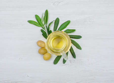 Green olives and olive oil in a glass jar with olive tree branch top view on a white wood plank backgroundの写真素材