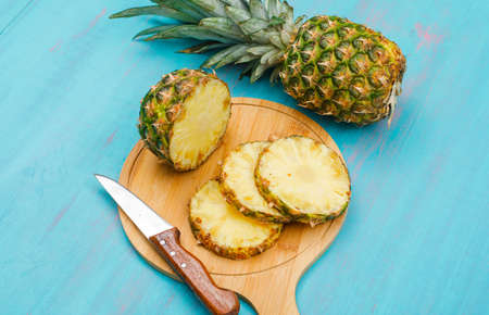 Whole and sliced pineapple with a knife in a cutting board on blue cyan background, high angle view.