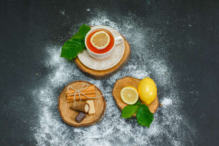 A cup of tea with lemon, leaves, dry cinnamon on wood slices and dark background, top view. 