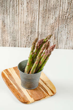Edible, raw stems of asparagus in a mini bucket with wooden board high angle view on a grunge wooden and white backgroundの写真素材