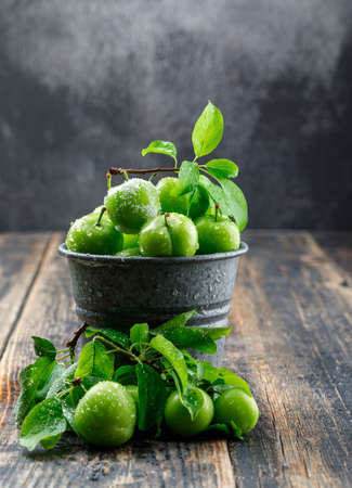 Cool green plums in a mini bucket with leaves side view on wooden and misty backgroundの写真素材