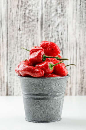 Red peppers in a mini bucket side view on white and wooden grunge backgroundの写真素材