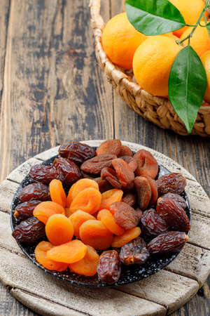 Dried apricots in a plate with dates and oranges in basket high angle view on wooden and cutting board backgroundの写真素材