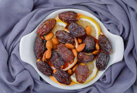 Dates and almonds with citrus fruit slices in a white plate on textile background, top view.