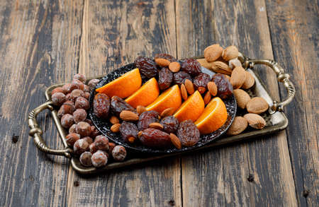 Nuts mix with dates and sliced orange in tray and plate on wooden background, high angle view.の写真素材