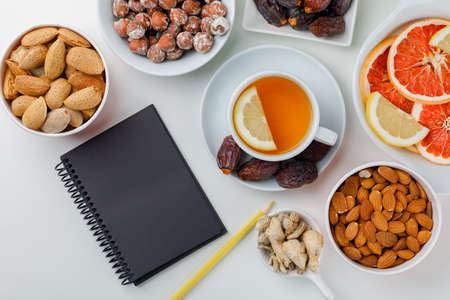 Dates, almonds, nuts in white plates with lemony tea, ginger, citrus fruits, pencil and notebook flat lay on a white backgroundの写真素材