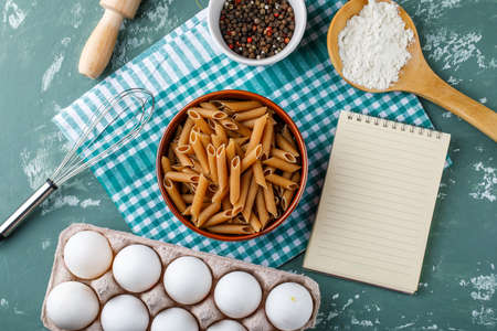 Penne pasta with eggs, peppercorns, starch, whisk, rolling pin, copybook in a bowl on grunge and kitchen towel background, flat lay.の写真素材