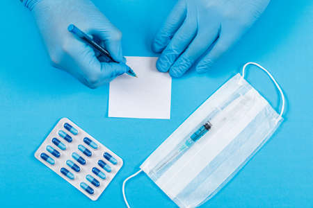 Medical concept with sticky note, pack of capsules, face mask, injection, sticky note on blue desk backgroud flat lay. doctor writing with pen.の写真素材