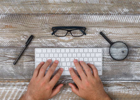 Top view a man typing on keyboard with eyeglasses, pen, magnifier on wooden background. horizontal