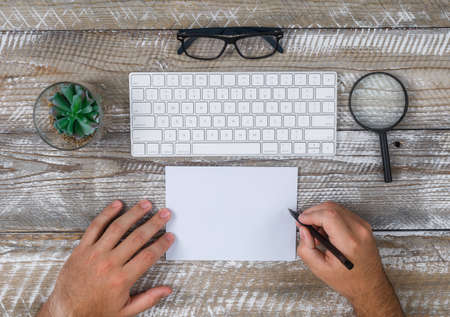 Top view man writing something on paper, with eyeglasses, pen, keyboard, cactus, magnifier on wooden background. horizontalの写真素材