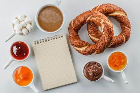 Flat lay notepad and a cup of coffee with jams, raspberry, sugar, chocolate in cups, turkish bagel...