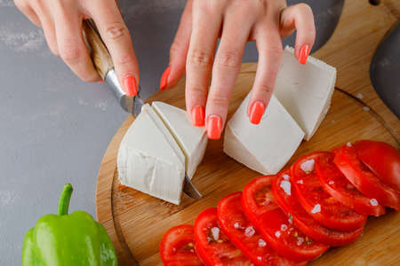 Some woman slicing cheese with sliced tomatoes, on a cutting board on gray background, high angle...