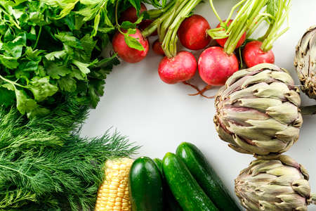 Artichokes and radishes with cucumbers, corn, greenery flat lay on a white background