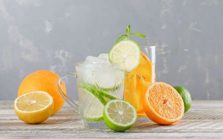 Detox water with limes, lemons, oranges, mint in cup and glass on wooden and plaster background,...
