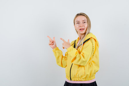 blonde girl pointing left with index fingers in pink t-shirt and yellow jacket and looking beautiful. front view.の写真素材
