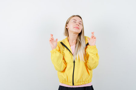 blonde girl in pink t-shirt and yellow jacket holding fingers crossed and looking beautiful , front view.の写真素材