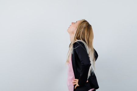 blonde girl holding hands on waist, looking above in pink t-shirt and black jacket and looking serious , front view.の写真素材