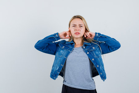 young lady in t-shirt, denim jacket, skirt plugging ears with fingers and looking confused , front view.の写真素材