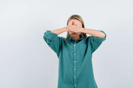 blonde woman holding hands on eyes in green shirt and looking scared , front view.の写真素材