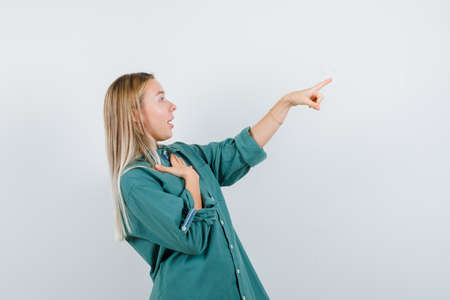 blonde girl in green blouse holding hand on chest while pointing right and looking surprised , front view.の写真素材