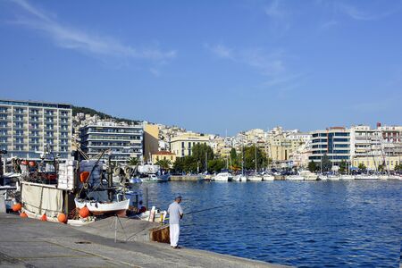 Kavala, Greece - September 17th 2015: Unidentified man fishing in the harbor, homes and fishing boatのeditorial素材