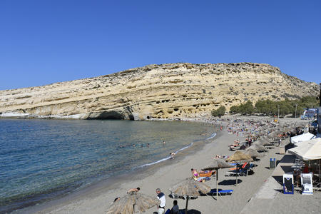 Matala, Greece - October 07, 2018: Unidentified people on sandy beach in the village in the south of Crete on Libyan sea with ancient tombs on hillのeditorial素材