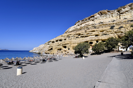 Matala, Greece - October 07, 2018: Unidentified people on sandy beach with ancient tombs on right and Paximadia islands left on backgroundのeditorial素材