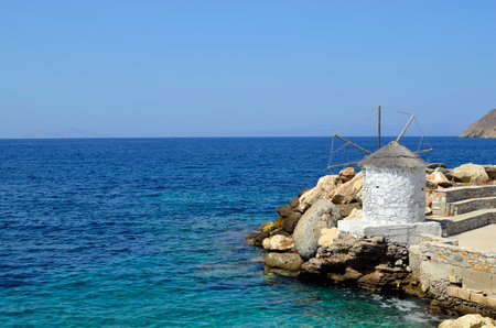 View of a small windmill at the jetty in the port of Aigiali with the clear blue Aegean Sea in the background.の写真素材