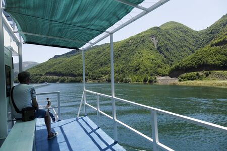 Koman, Albania, July 7 2019: A tourist sits on the railing and enjoys the scenery on the Komani lake in the dinaric alps of Albaniaのeditorial素材