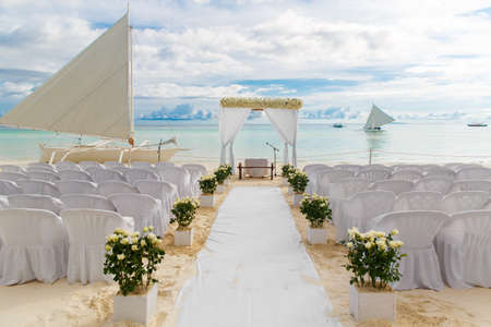 Wedding ceremony on a tropical beach in white. The arch is decorated with flowers on the sandy beach, decorated chairs for guests, sand castle and yacht in the sea. Wedding and honeymoon concept.の写真素材