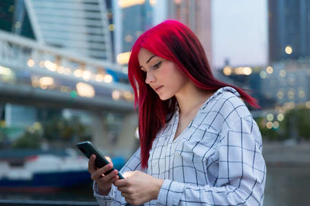 Beautiful red-haired girl with smartphone in the evening on lighted city street.の写真素材