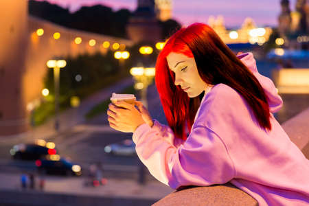 Beautiful red-haired teenage girl in pink hoodie drinking coffee in the evening on lighted city...