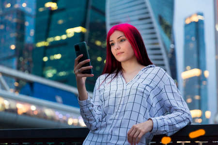Beautiful red-haired girl with smartphone in the evening on lighted city streetの写真素材
