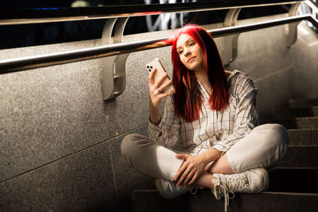 Beautiful red-haired teenage girl with smartphone sitting on the steps on street night-lit city.の写真素材