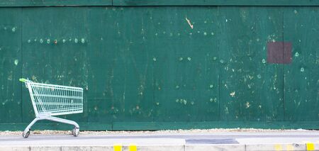 Green fence background. Copy space next to supermarket cart on the pavement.