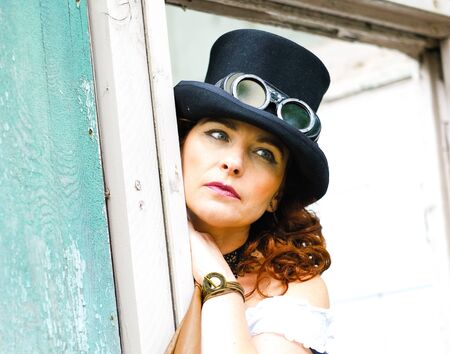 Beautiful redhair steampunk girl with goggles on black hat outside toilet background.