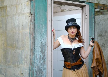 Beautiful redhair steampunk girl with goggles on black hat outside toilet background.